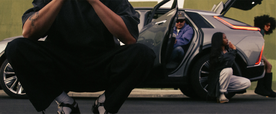 Close-up of Oakland Artist Esteban Samayoa Kneeling and Posing in Front of Three Friends Sitting Inside a Cadillac LYRIQ