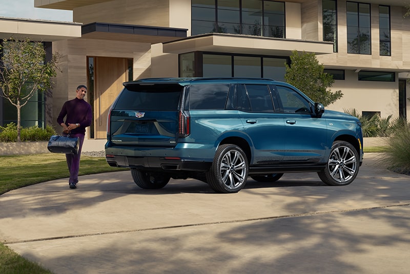 Three Quarters View of a Blue Cadillac SUV Parked in Front of a House While a Man Walks to the Trunk with a Bag